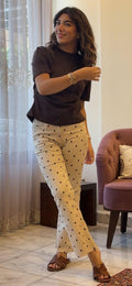 A woman modeling cream-colored pants with a black polka-dot pattern, paired with a dark brown t-shirt and brown sandals, standing in a room with a window, sheer curtains, and a purple armchair.