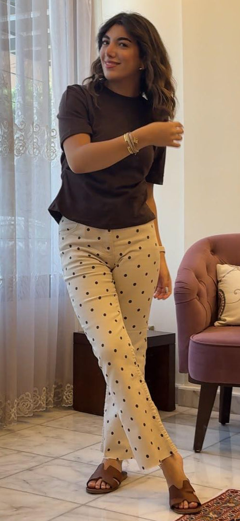 A woman modeling cream-colored pants with a black polka-dot pattern, paired with a dark brown t-shirt and brown sandals, standing in a room with a window, sheer curtains, and a purple armchair.